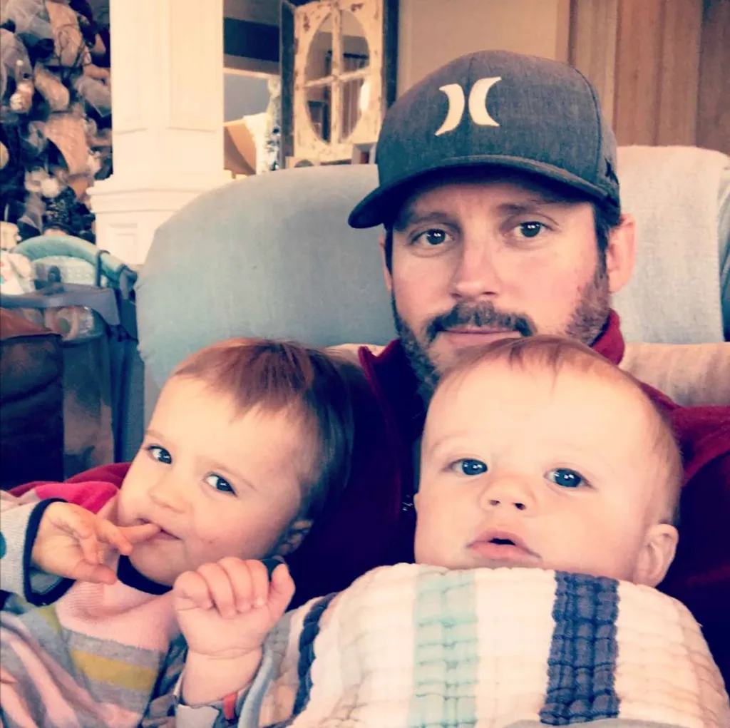 Brandon Blackstock, Kelly Clarkson's ex-husband, wearing a baseball cap and holding their two young children.
