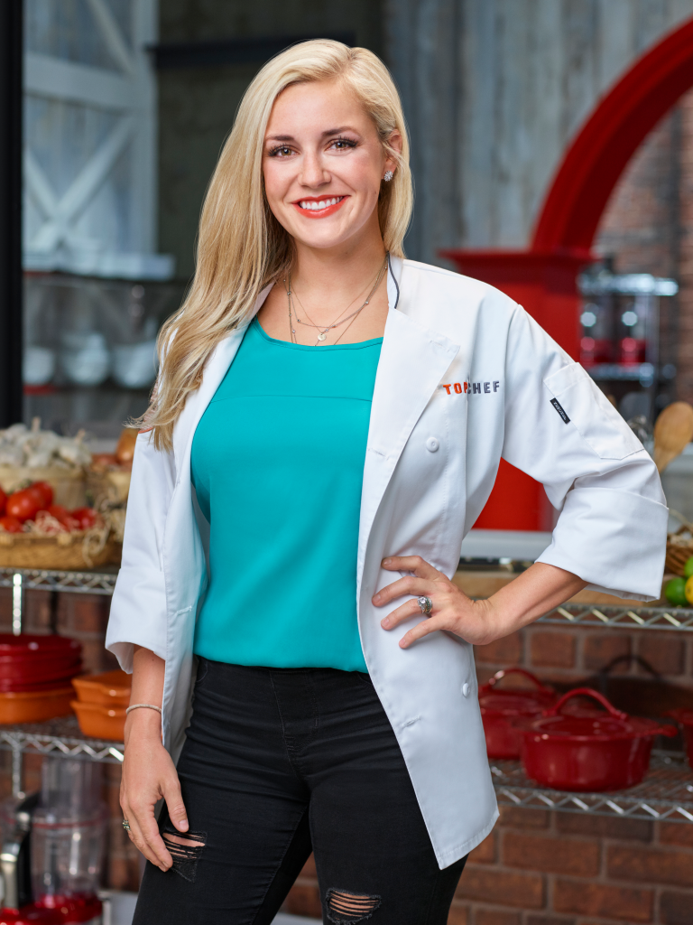 Kelsey Barnard Clark, a blonde woman wearing a white chef coat over a teal shirt and black distressed jeans, smiles at the camera.