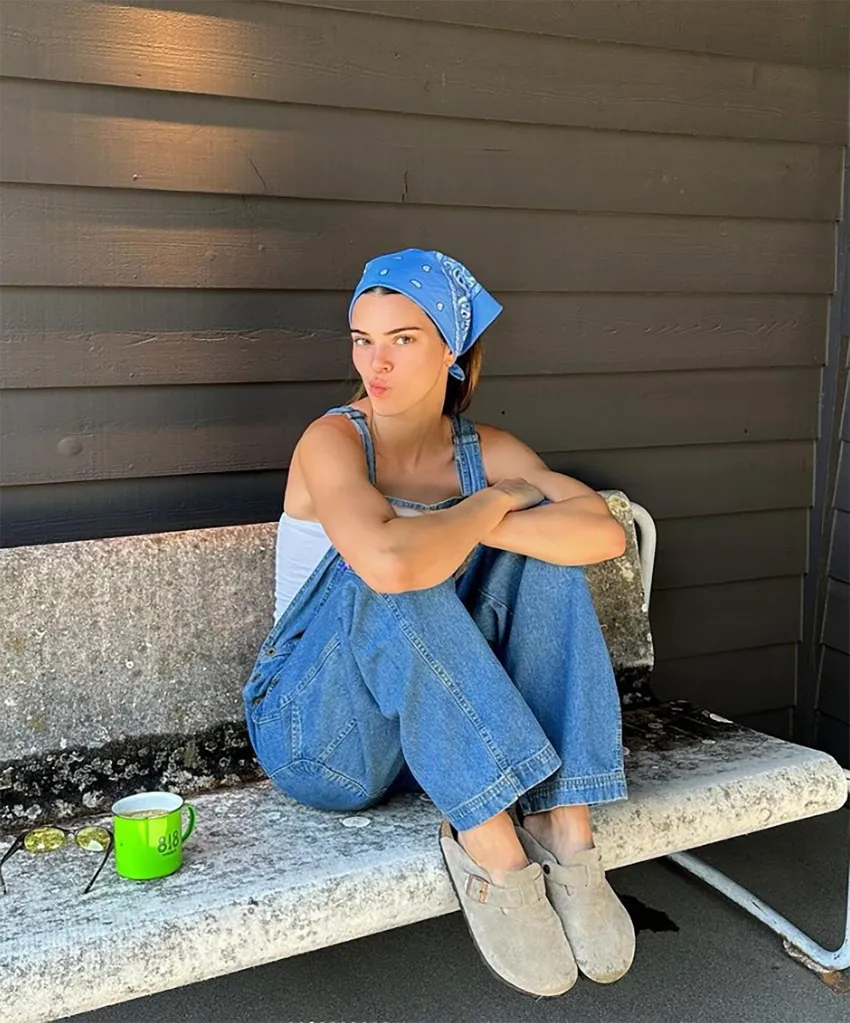 Kendall Jenner sitting on a bench, wearing a blue bandana, white tank top, and denim overalls.