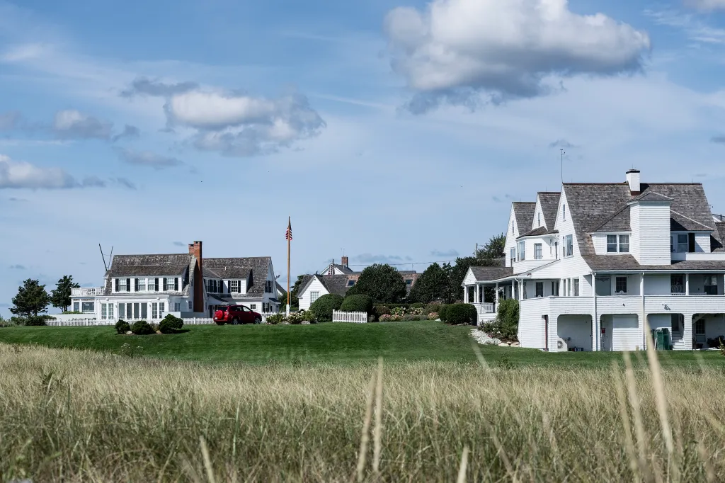 Exterior view of the Kennedy Compound in Hyannis Port, Massachusetts, with the main house on the left, a red car, and another house on the right, all behind a grassy lawn and tall grass in the foreground.