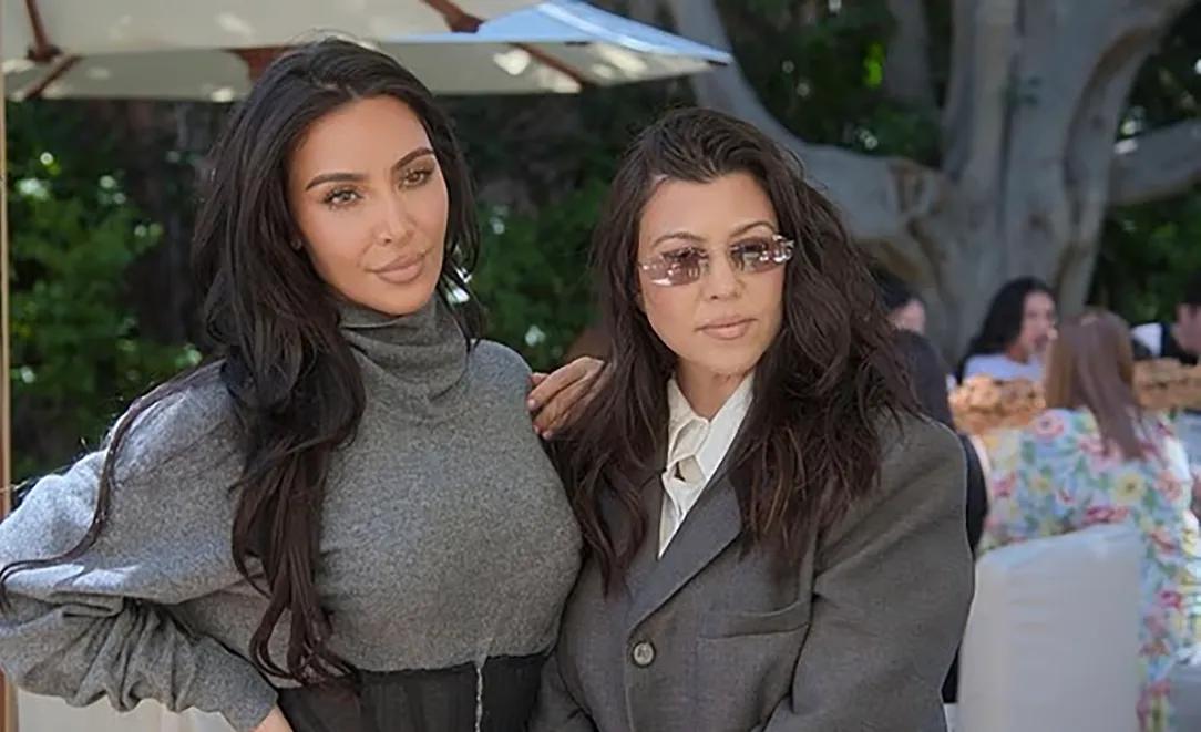 Kim Kardashian in a gray turtleneck and Kourtney Kardashian in a gray blazer and sunglasses.