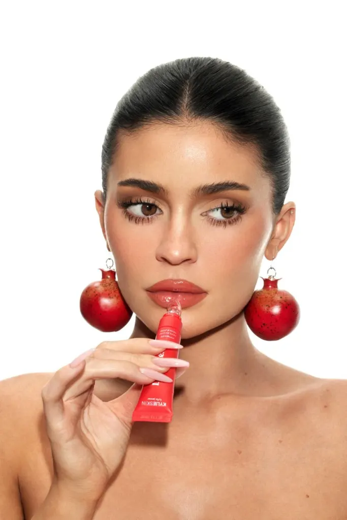 Kylie Jenner applying lip gloss while wearing pomegranate earrings.