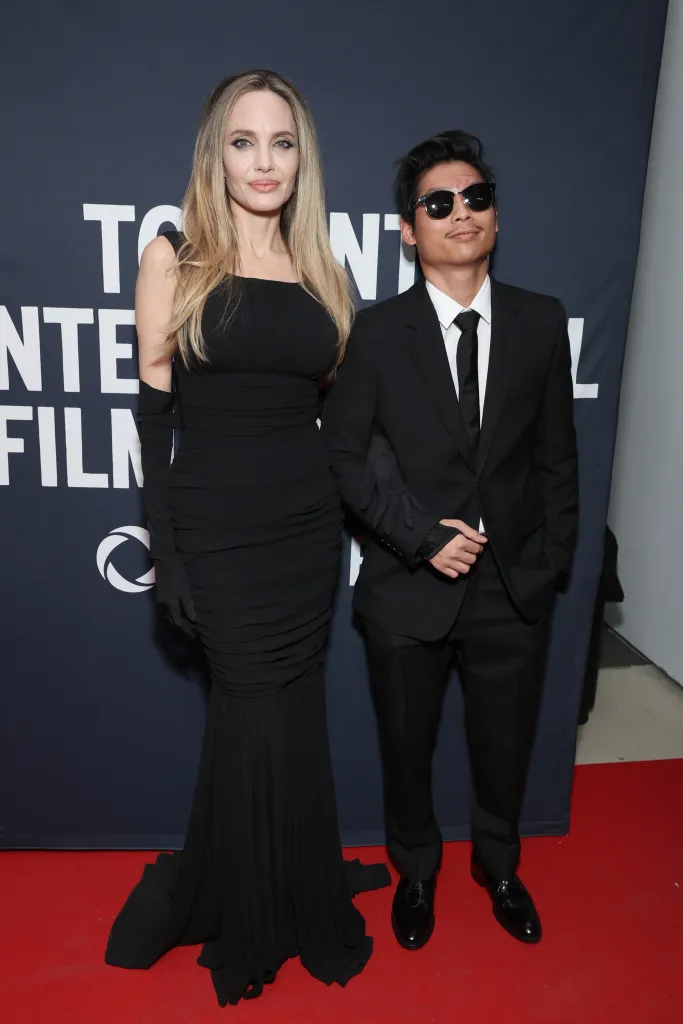 Angelina Jolie in a long black gown and long gloves, arm in arm with Pax Jolie-Pitt in a black suit and sunglasses, posing on a red carpet.