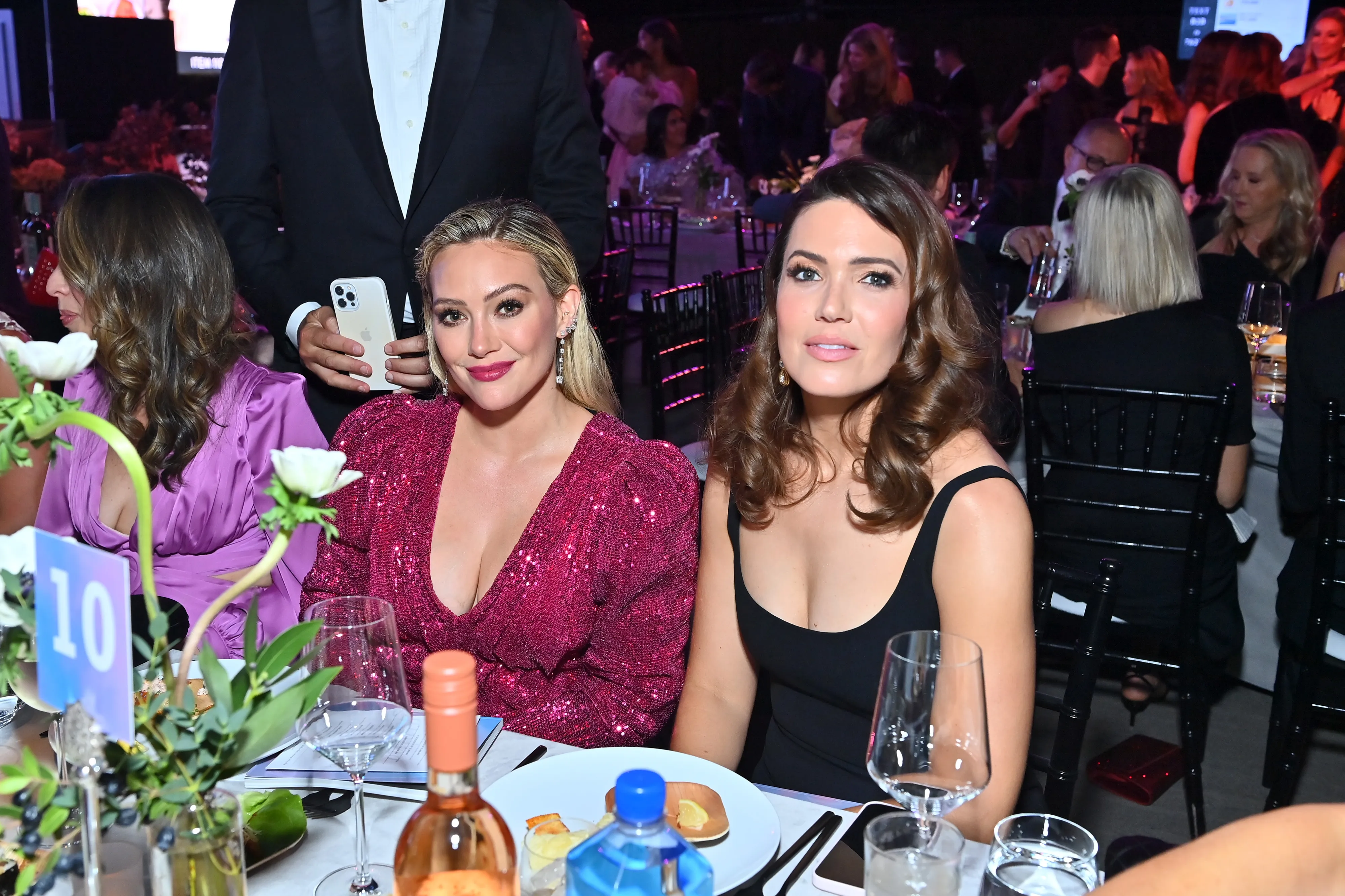 Hilary Duff in a pink sequined dress and Mandy Moore in a black dress sit next to each other at a table.