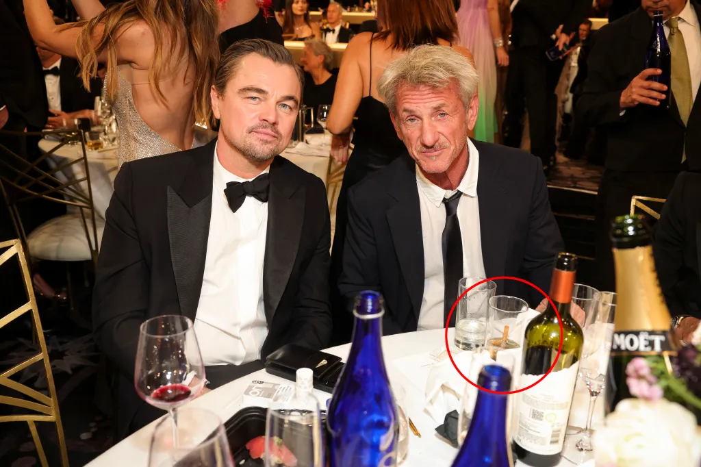 Leonardo DiCaprio and Sean Penn sitting at a table with drinks and what appears to be a cigarette in a glass.