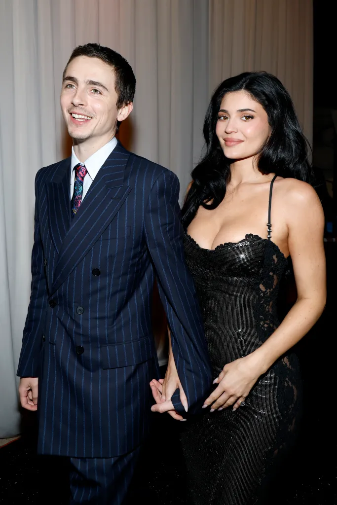 Timothée Chalamet in a pinstriped suit and Kylie Jenner in a black shimmering dress hold hands.