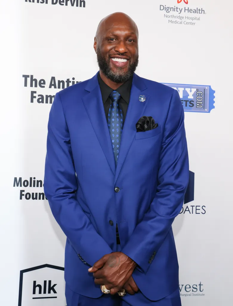 Lamar Odom smiling while wearing a blue suit, black shirt, and blue patterned tie.