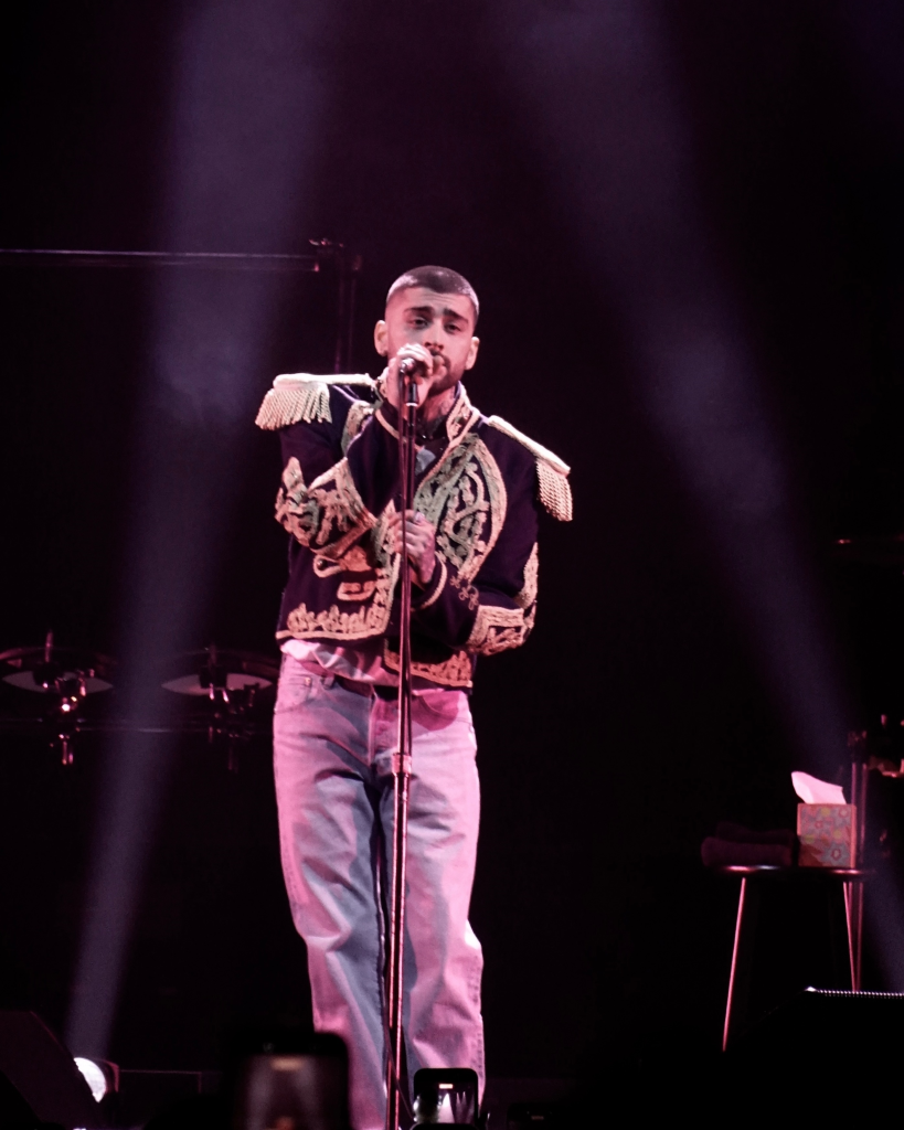Zayn Malik performing on stage in a navy jacket with gold embroidery and light blue jeans.