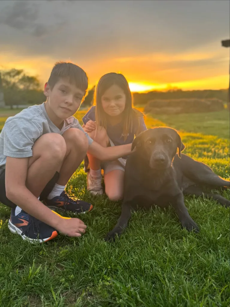 A boy and a girl squatting in a field with a black dog at sunset.