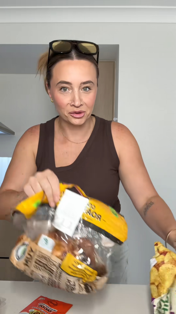 A woman holds a bag of rotisserie chicken in one hand and a bag of tortilla chips in the other, ready to prepare a meal.