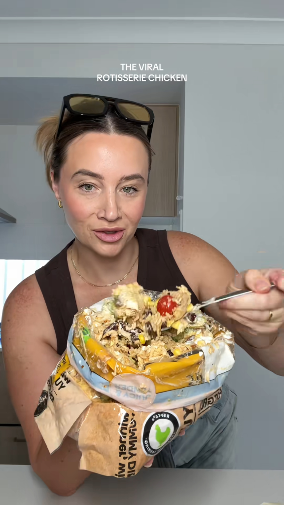 A woman holds up a bag of rotisserie chicken mixed with salad ingredients, serving herself with a spoon.