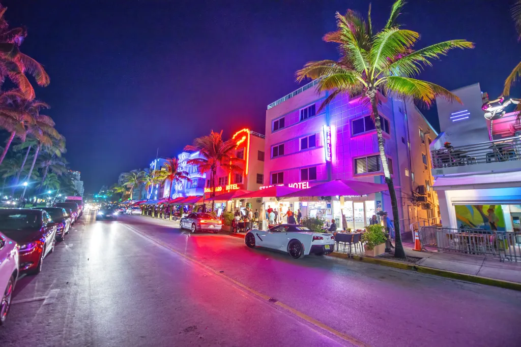 Ocean Boulevard at night with palm trees and illuminated buildings housing restaurants with outdoor seating, cars parked and driving.