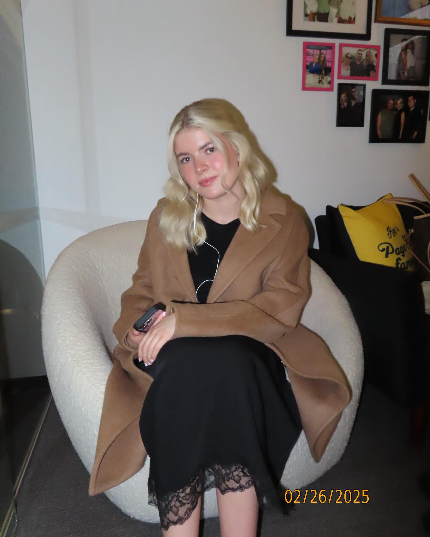 Lola Sheen seated in a chair, wearing a black dress and a light brown coat, holding a phone.
