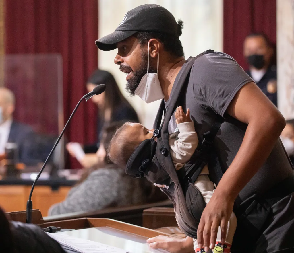 Jason Reedy speaking at a Los Angeles City Council meeting while holding a baby in a carrier.