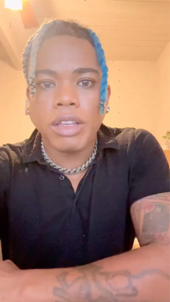 A person with braided blue and grey hair and a thick cuban link chain around their neck is looking at the camera.