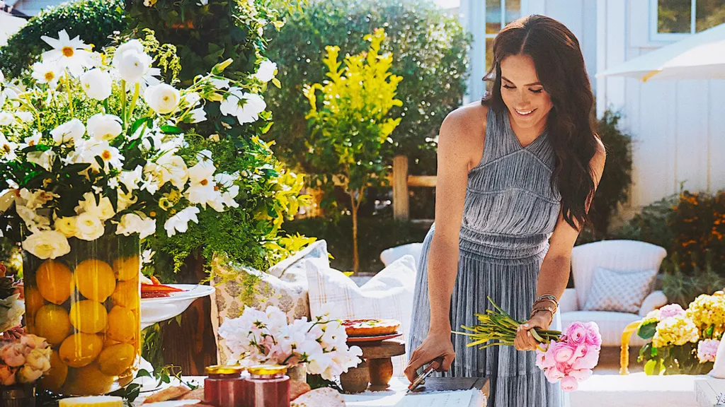Meghan Markle, in a grey summer dress, arranging flowers on a table in an outdoor setting.