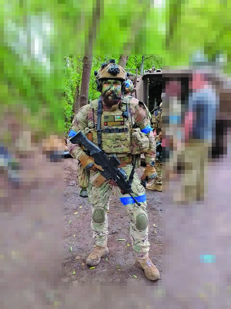 A soldier in camouflage, wearing face paint, helmet, and tactical gear, holding a rifle with an M113 personnel carrier visible behind him.