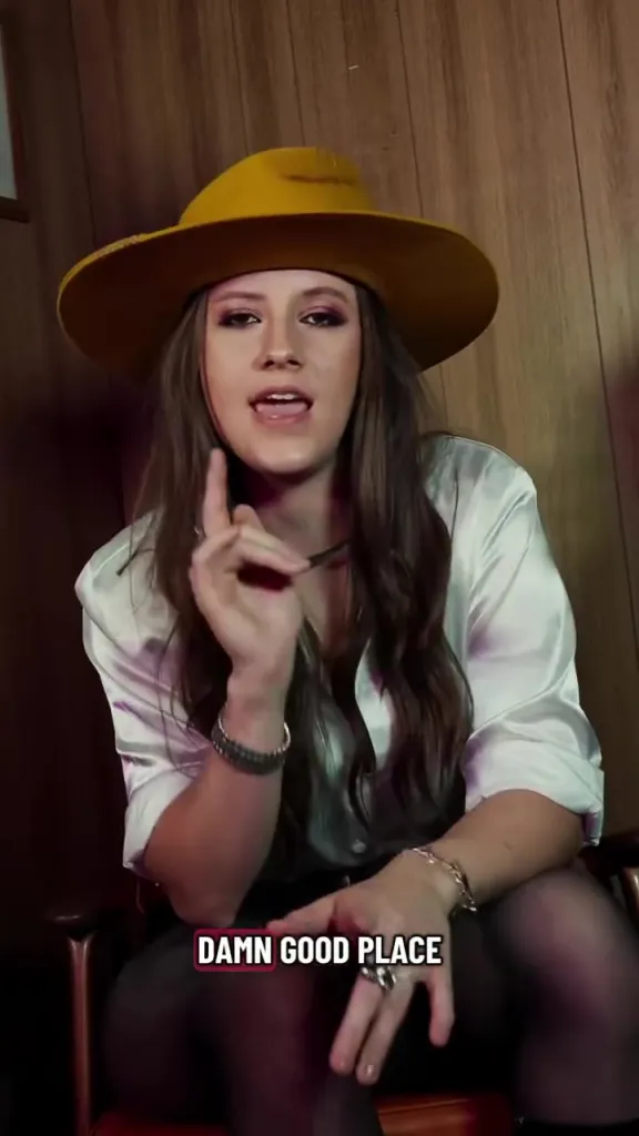 Maggie Baugh singing about trying to fight feelings, wearing a yellow hat, white satin shirt, and a black necklace.