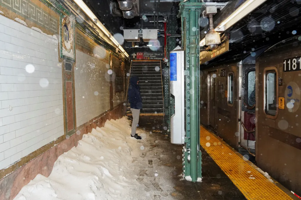A man waits to board an arriving train as snow falls inside the Borough Hall subway station.