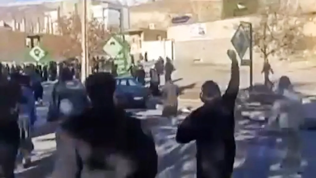 A blurry image showing people protesting in the Malekshahi district of Iran's western Ilam province, with one person in the foreground raising their fist.