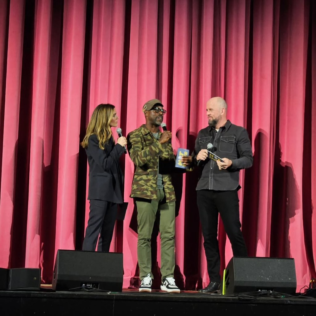 Mandy Moore, her husband Taylor Goldsmith, and an unidentified man holding a microphone stand on stage in front of a red curtain.