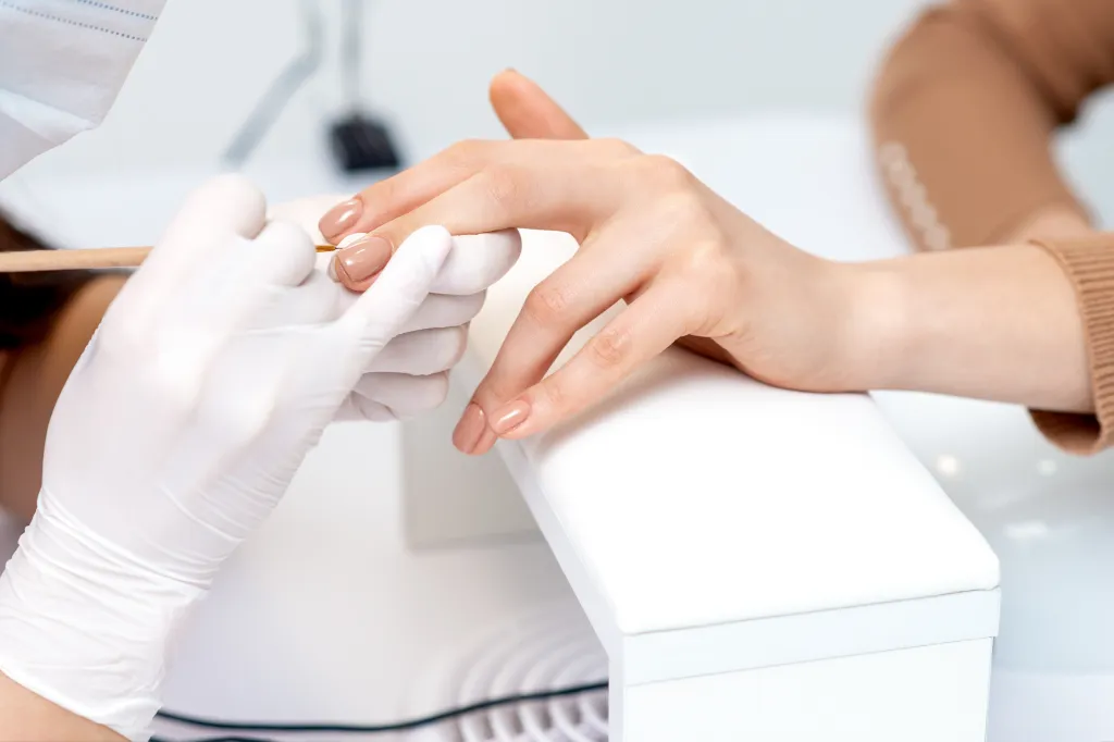 A close-up of a manicurist in white gloves applying beige nail polish to a client's fingernail.