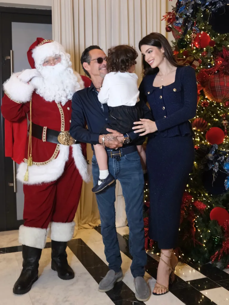 Marc Anthony holding a child, standing next to Nadia Ferreira, with Santa Claus beside them in front of a Christmas tree.