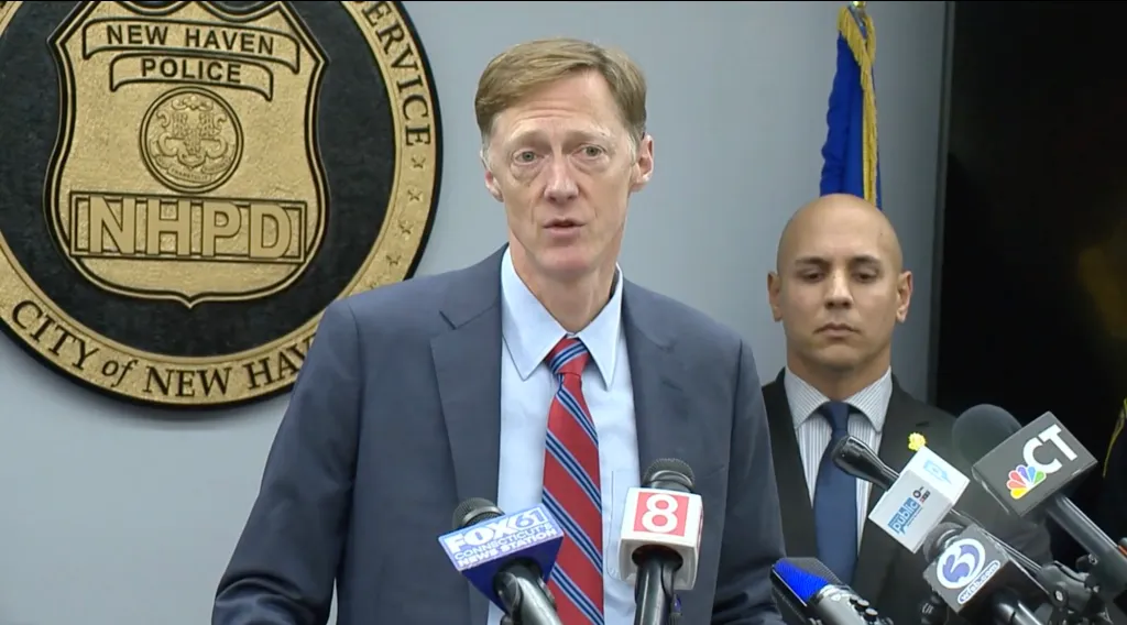 New Haven Mayor Justin Elicker speaking at a press conference with news microphones in front of him.