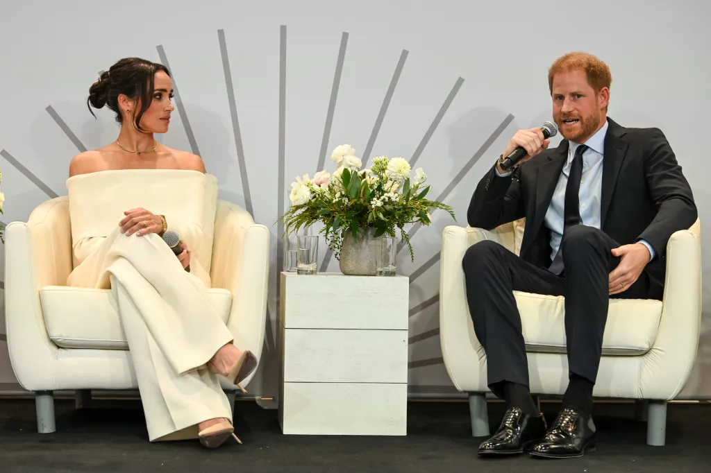Meghan Markle, in a white off the shoulder top and white pants, and Prince Harry, in a grey suit, blue shirt and black tie, speaking at a mental wellness event.