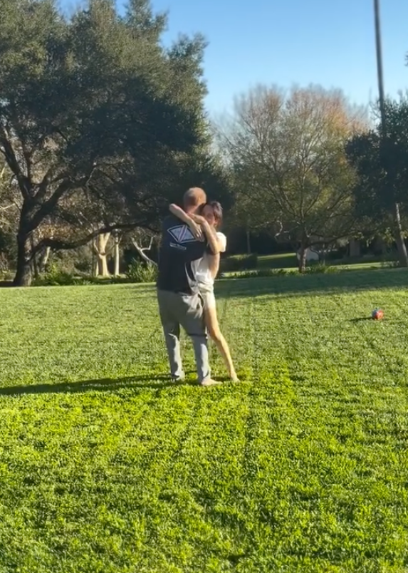 Meghan Markle, in.a white t-shirt and cream shorts, and Prince Harry, n a blue t-shirt and khaki pants, dancing on a grassy lawn.
