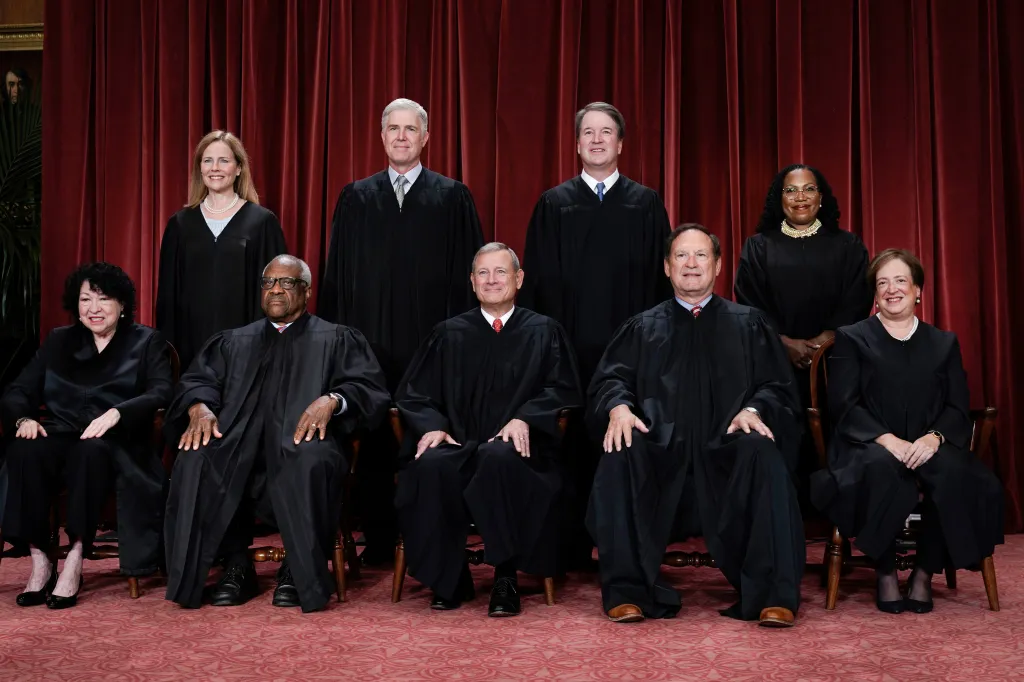 Nine Supreme Court Justices pose for a group portrait, five seated in front and four standing behind, all wearing black judicial robes.