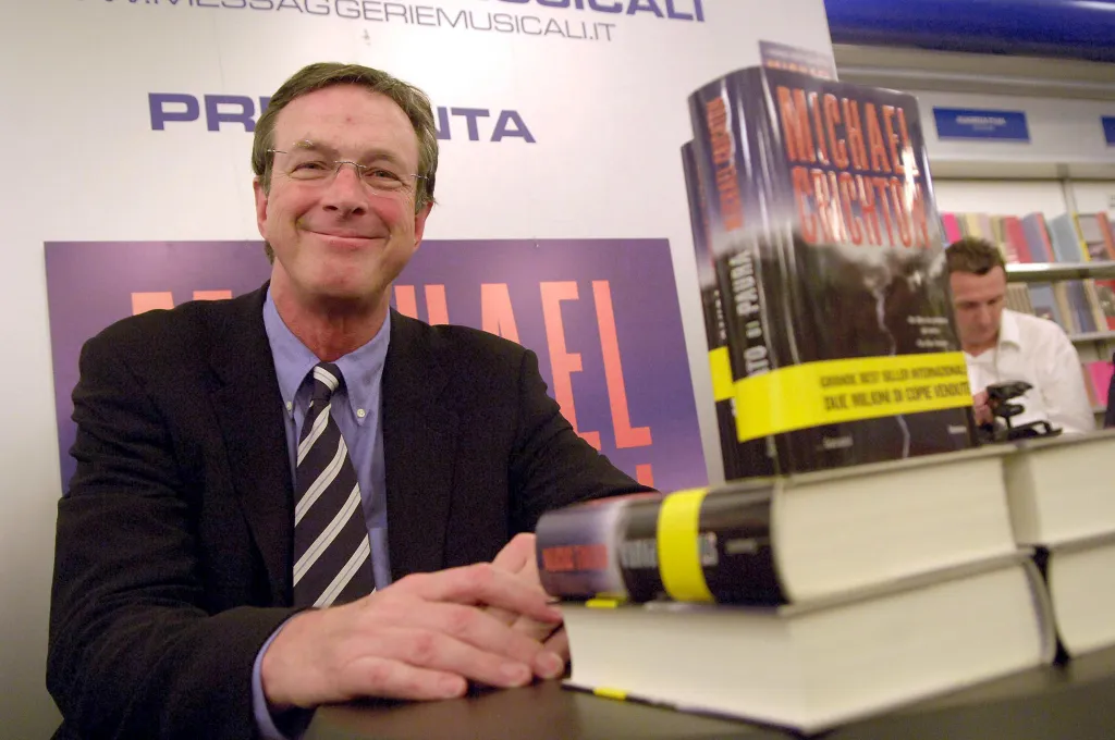 Michael Crichton, in a black suit, blue shirt and striped tie, smiling at his book launch, with his books prominently displayed.