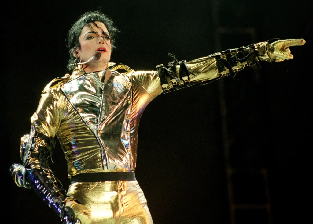 Michael Jackson performing in a shimmering gold outfit with a microphone headset, pointing his arm and finger to the right.
