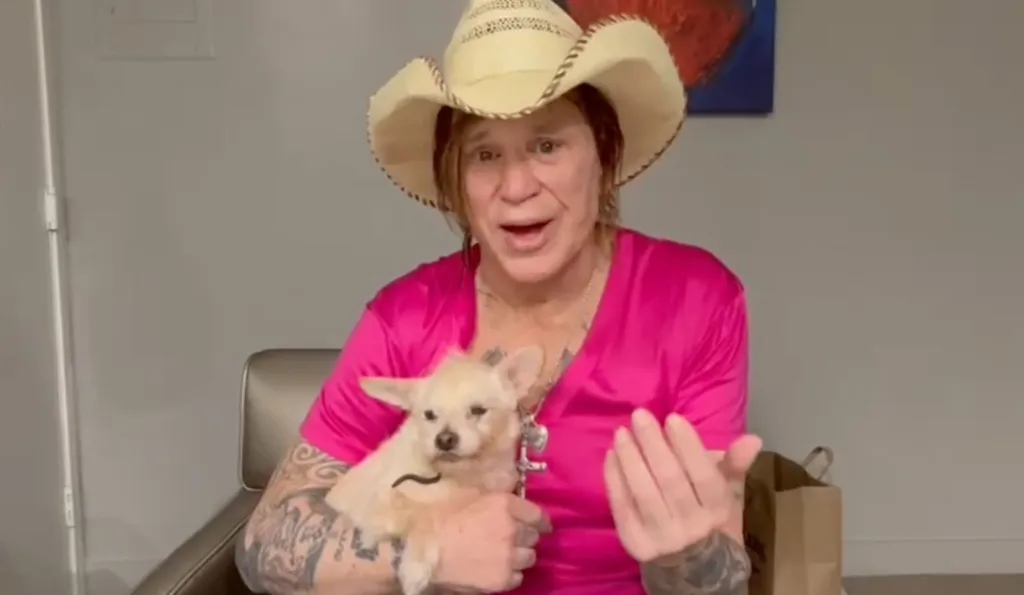 Mickey Rourke in a cowboy hat and pink shirt holding a small, light-colored dog.