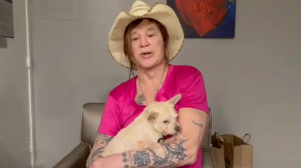 Mickey Rourke in a cowboy hat and pink shirt holding a dog.