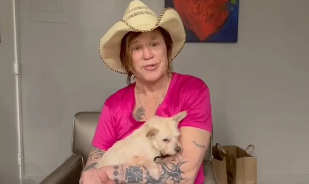 Mickey Rourke in a cowboy hat and pink shirt holding a dog.