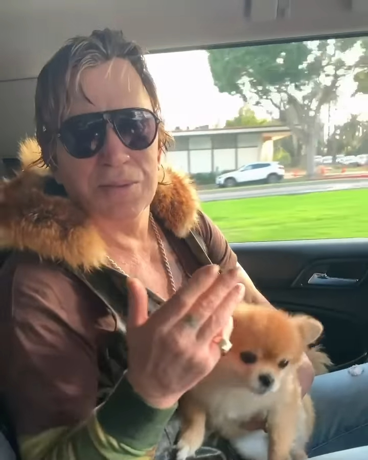 Mickey Rourke holding a Pomeranian dog while seated in a car.