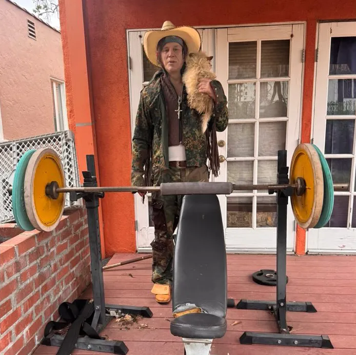 Mickey Rourke holding a small dog while standing next to a weight bench.