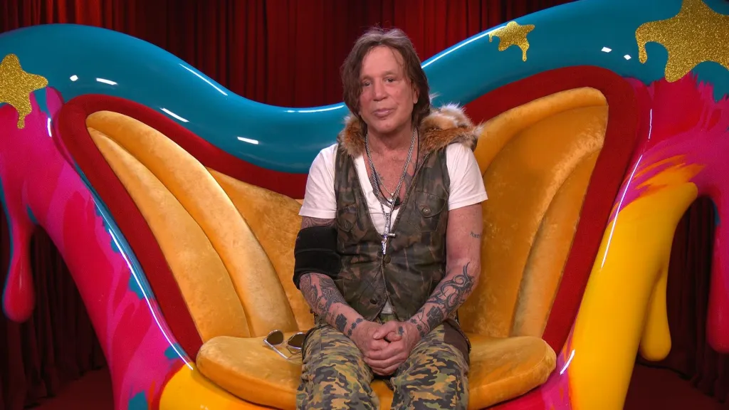 Mickey Rourke sitting in a colorful, oversized chair in the Big Brother diary room.