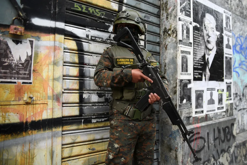A military police officer in camouflage uniform and helmet stands guard with a rifle, while posters are plastered on the wall behind him.