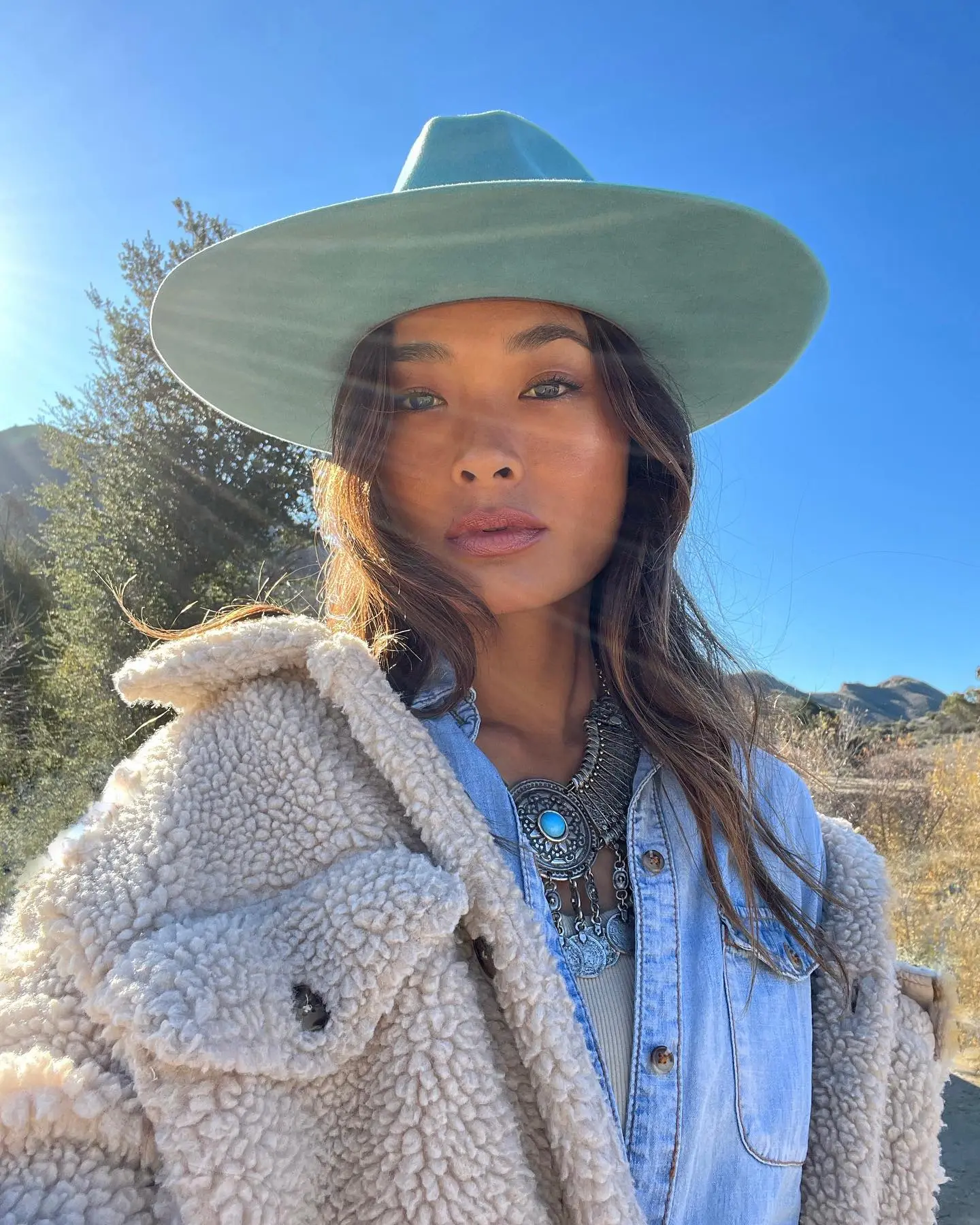 Jarah Mariano wearing a teal wide-brim hat, a large silver and turquoise necklace, a denim shirt, and a cream-colored sherpa jacket.