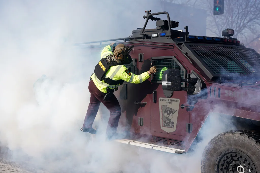 A Minnesota State Trooper in a gas mask and yellow high-visibility jacket holds onto an armored vehicle amidst smoke.