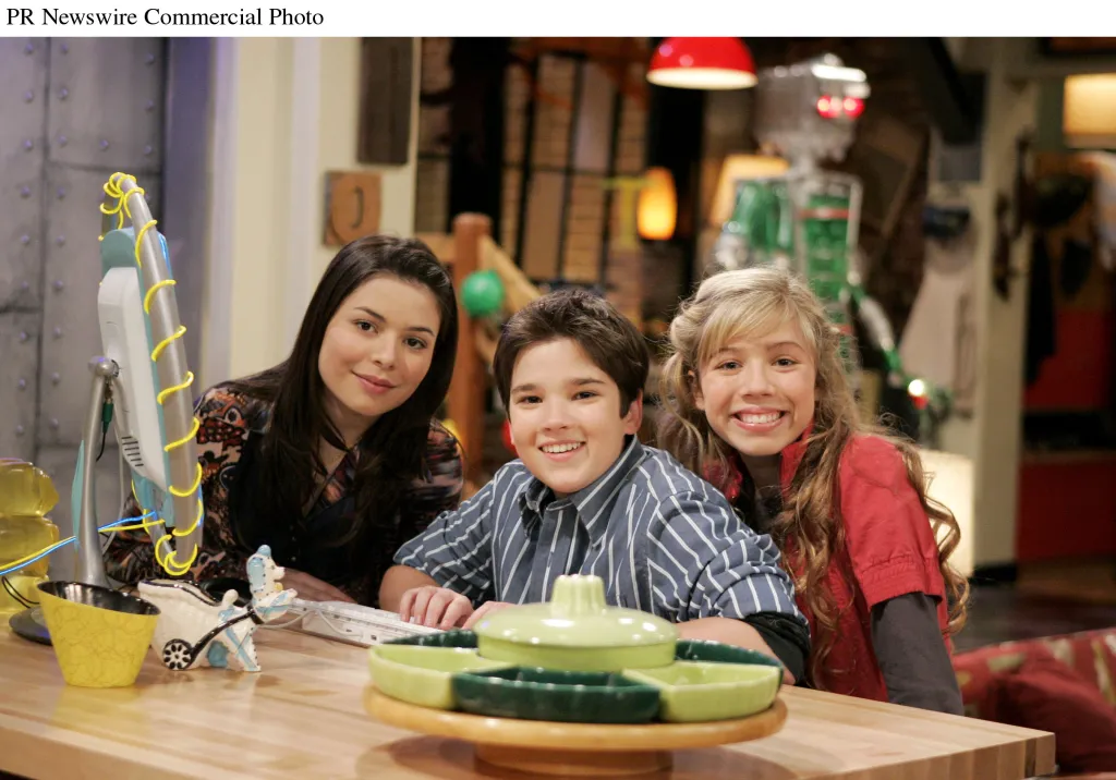 Miranda Cosgrove, Nathan Kress, and Jennette McCurdy smiling while sitting at a table with a computer monitor.