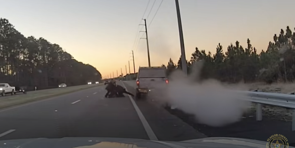 A person being apprehended on the side of a highway next to a stopped white truck with smoke coming from its exhaust.