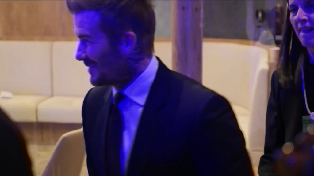David Beckham, in a suit, smiles and appears to try and ignore a nearby camera.