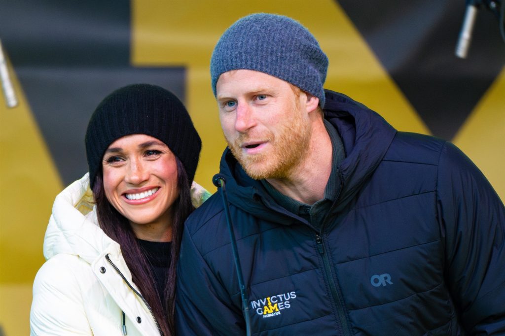 Prince Harry, Duke of Sussex, and Meghan Markle, Duchess of Sussex, dressed in winter attire with the Invictus Games Foundation logo visible on Harry's jacket.