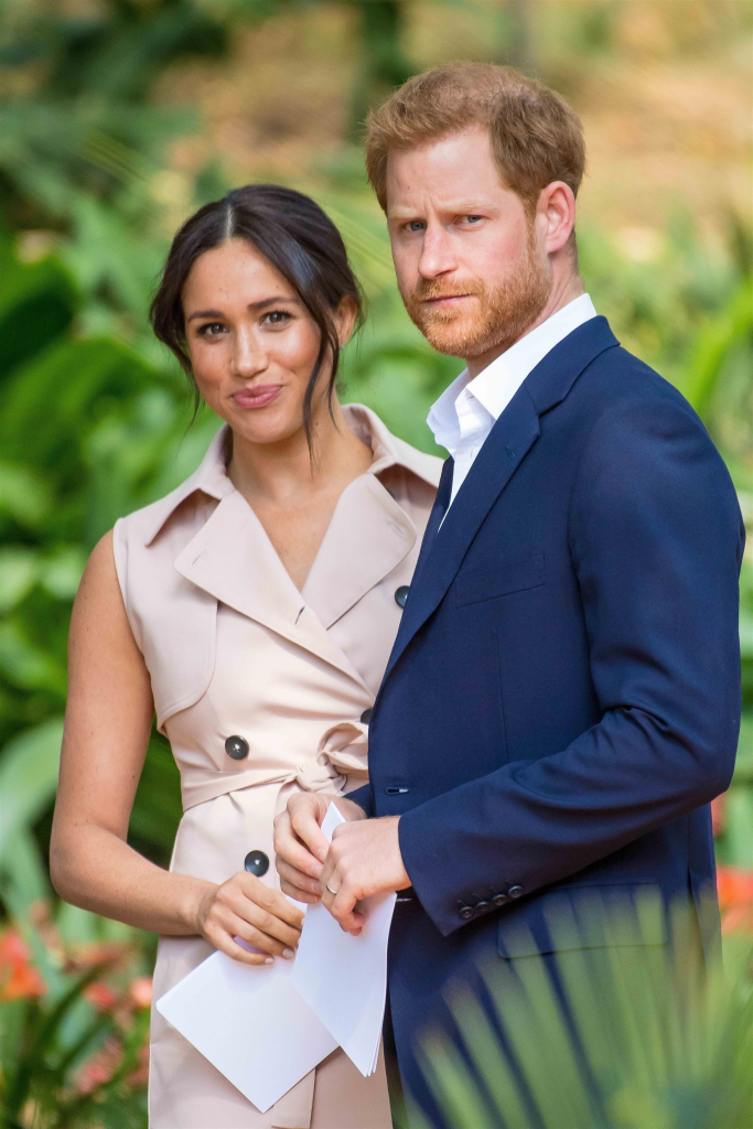 Meghan Markle, wearing a light-colored sleeveless dress, and Prince Harry, in a navy suit, holding papers, look forward at an event.