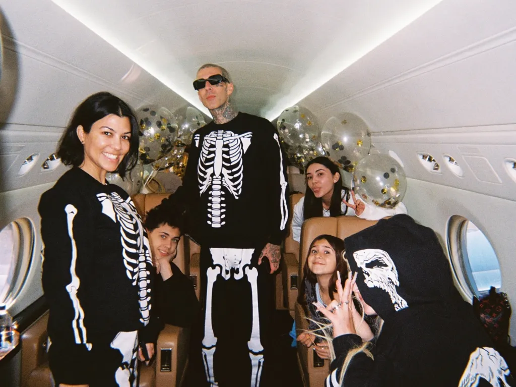 Kourtney Kardashian and Travis Barker with children Landon, Atiana, Penelope, and Alabama, all dressed in skeleton costumes, inside an airplane with balloons.