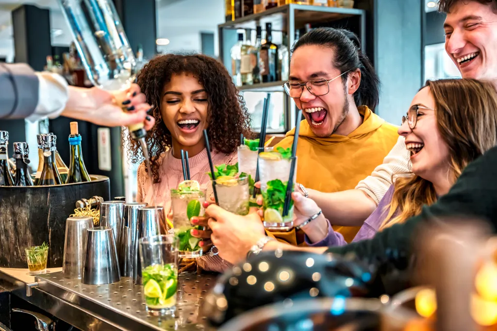 A group of friends laughing and cheering mojitos at a bar, while a bartender pours a drink.