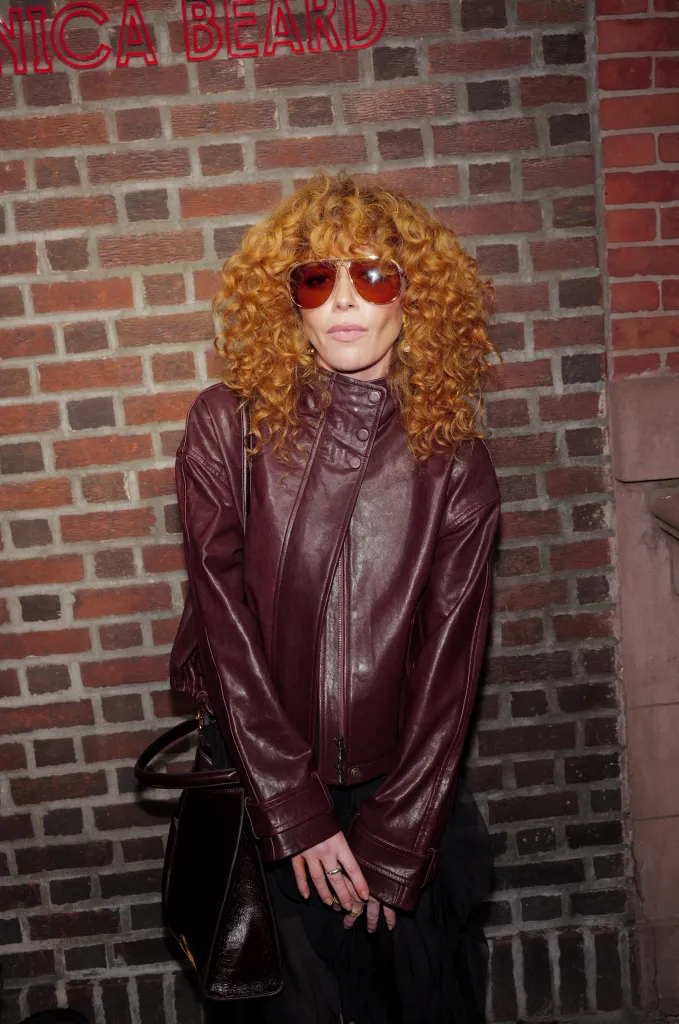 Natasha Lyonne in a maroon leather jacket and red sunglasses, standing in front of a brick wall.